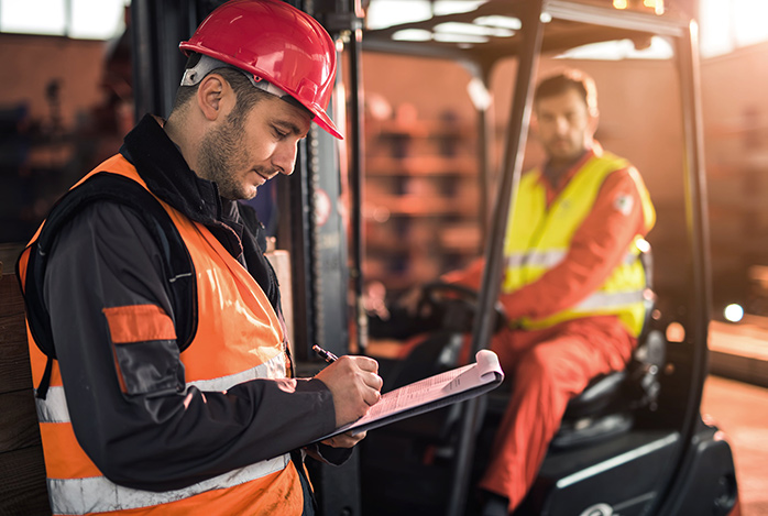 A health and safety officer making notes on a clipboard, with a forklift truck operator in the background