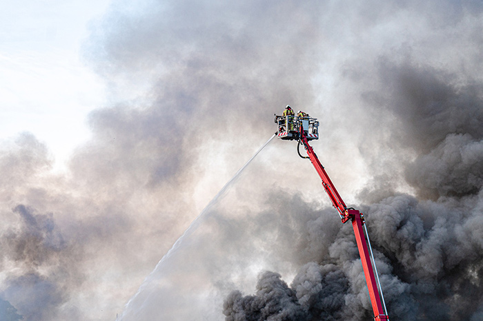 Two firemen in an elevated platform spraying water on a fire, with thick, black billowing smokestyle=